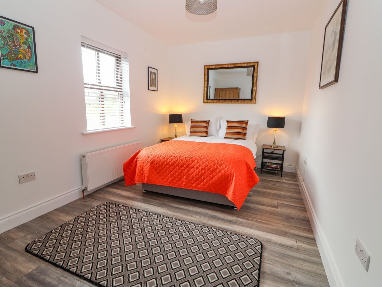 A bedroom with a bed, bedside lamps, a window with blinds, a radiator, and a patterned rug.
