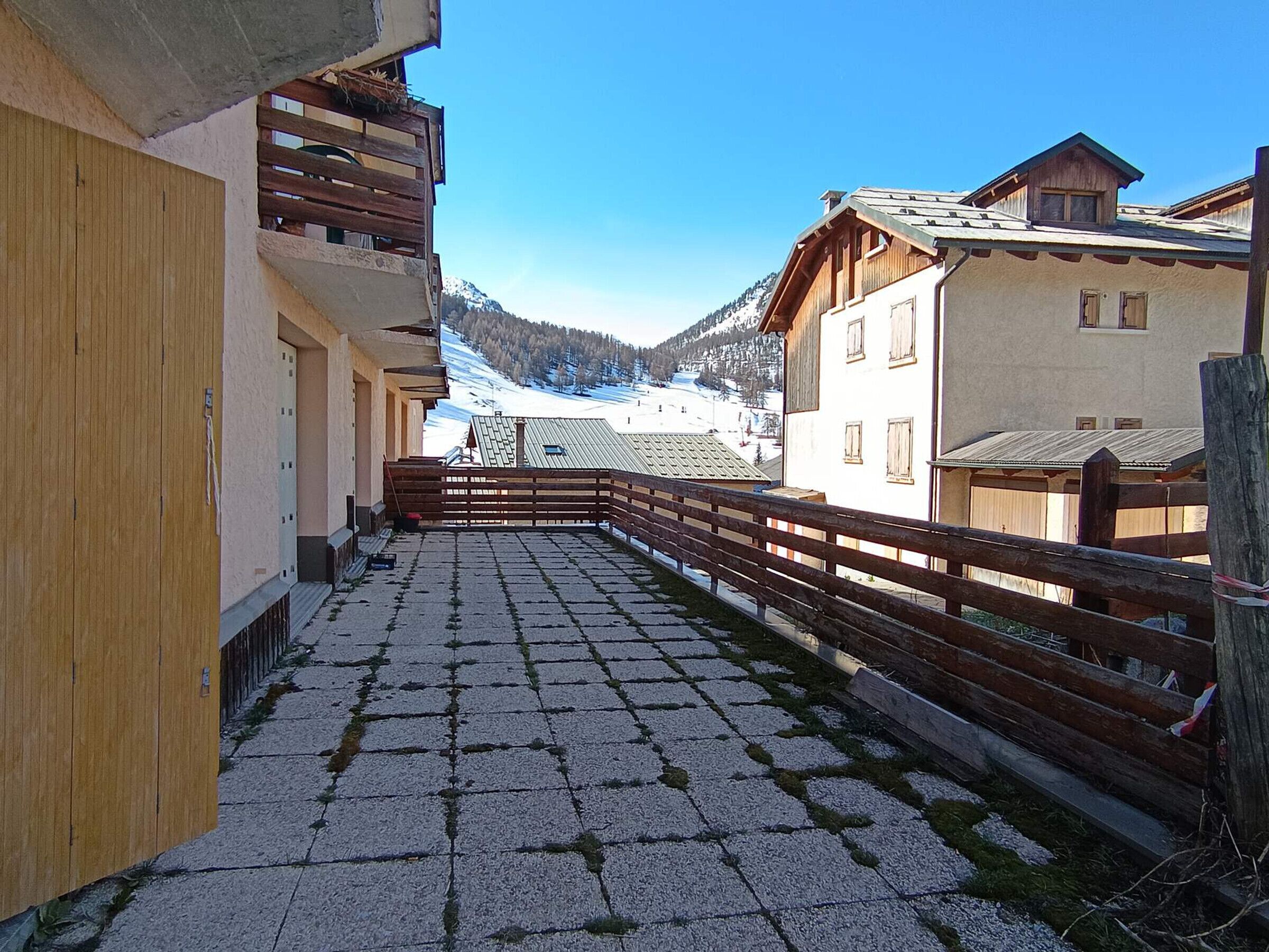 Une cour enneigée, entourée d’une clôture en bois, avec des bâtiments dotés de balcons et une vue sur les montagnes en arrière-plan.