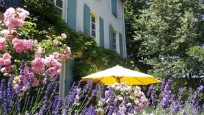 A building with a garden featuring pink and purple flowers, a yellow umbrella, and a tree in the background. - La Cerisaie (Riols)