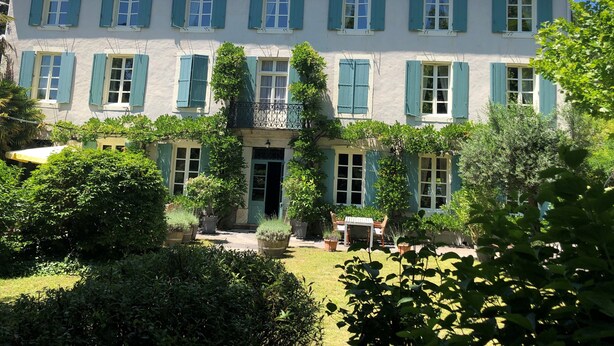 A two-story building with a tiled roof, multiple windows with shutters, and a well-maintained garden with potted plants and a seating area. - La Cerisaie (Riols)