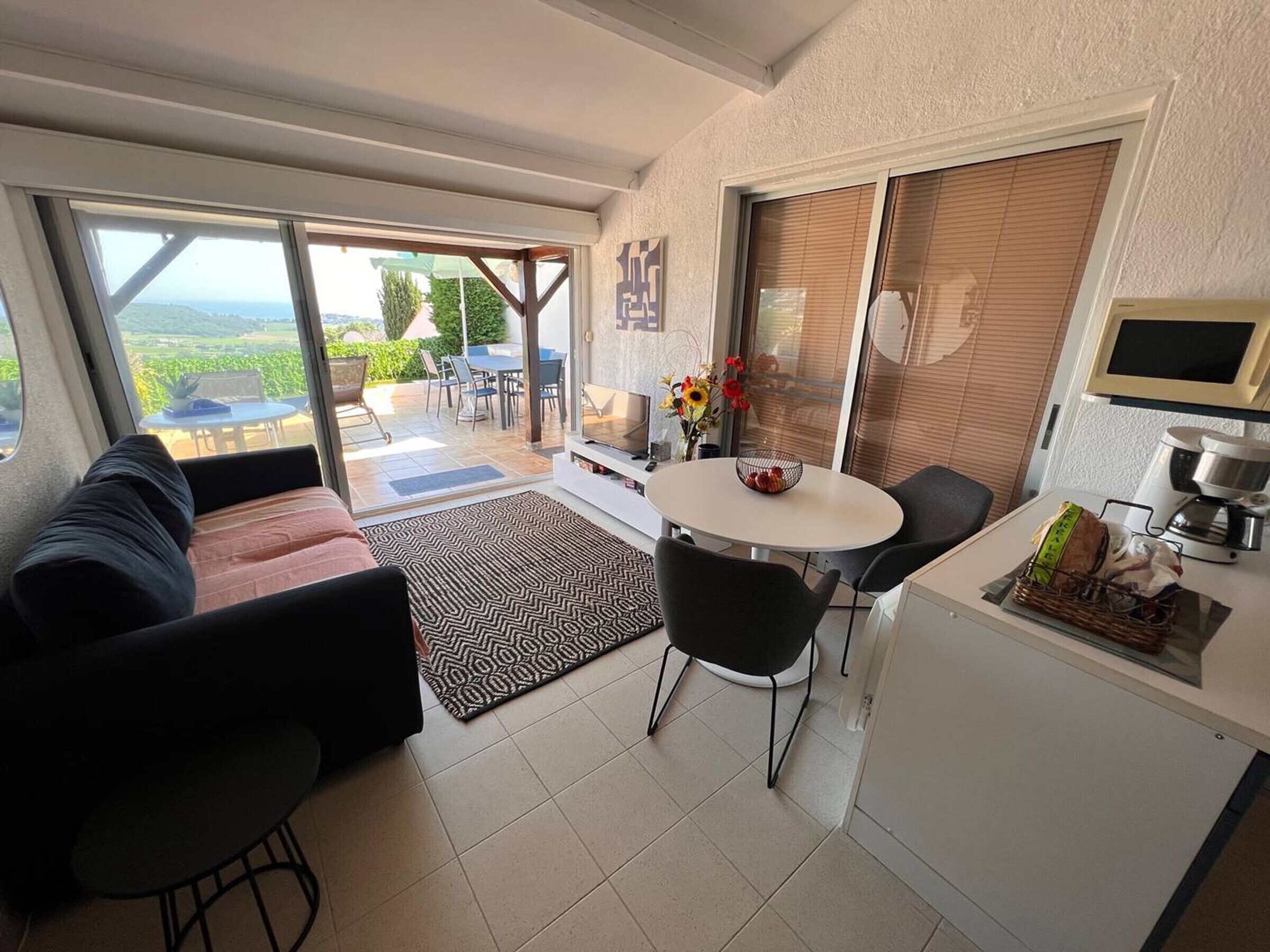 A modern living space with a sofa, a dining table with chairs, a coffee machine, and a view of a garden through large windows.