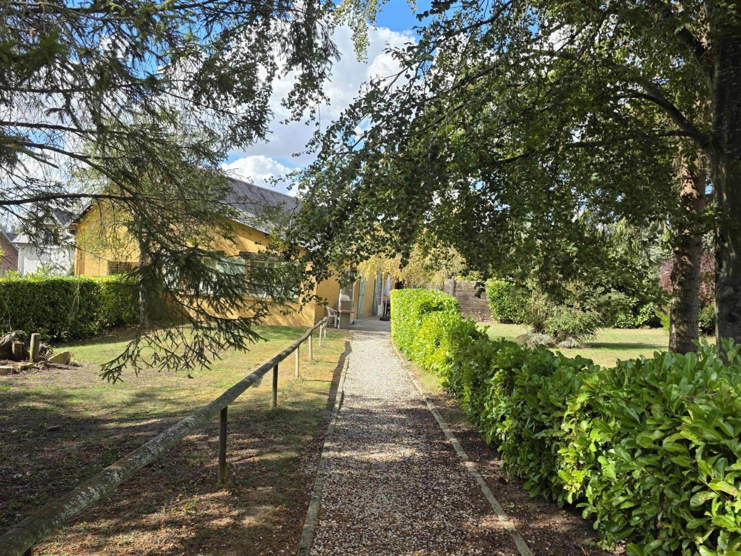 Un chemin de gravier mène à un bâtiment à la façade jaune, entouré de verdure et d’arbres.