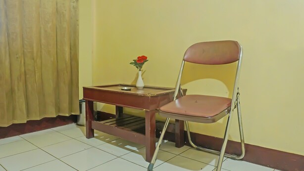 A small room with a chair, a table, and a vase with a red flower. - Hotel O Adam Kost SyariahNearWisata Jembatan Barito (Alalak)