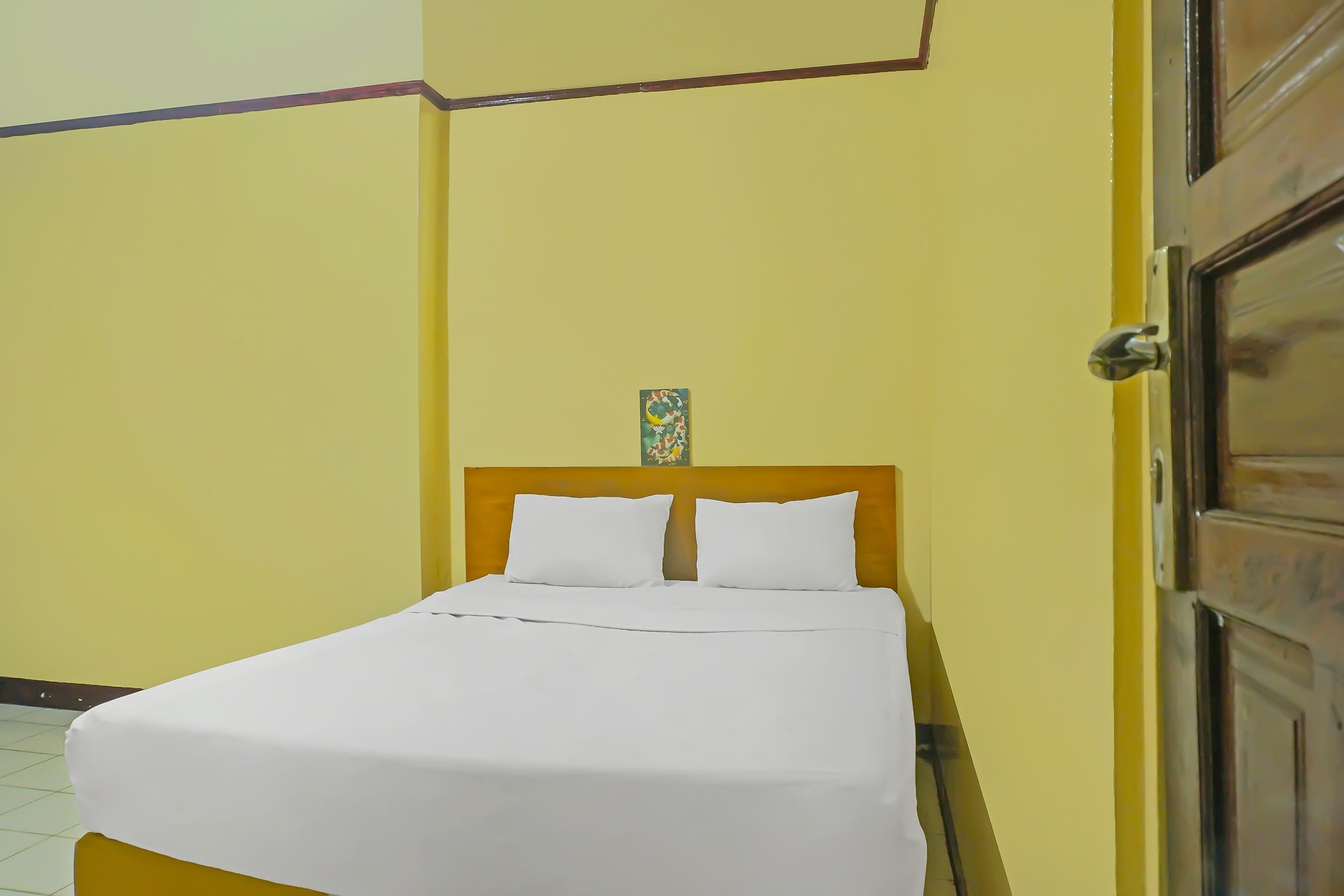 Un lit simple avec une literie blanche, dans une pièce aux murs jaunes et dotée d’une tête de lit en bois.