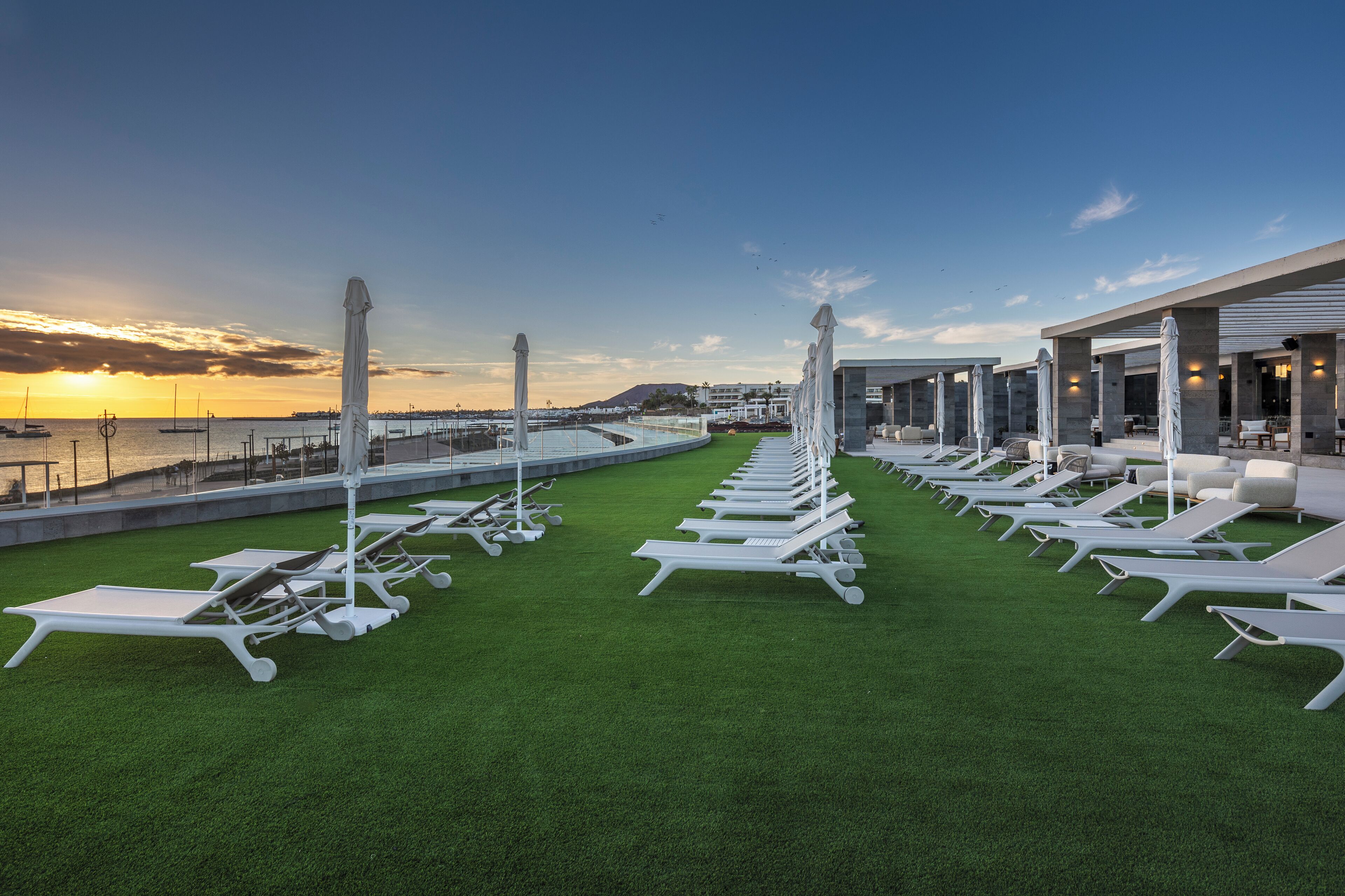 Zona de lounge en la azotea con filas de tumbonas blancas y sombrillas, con vistas a un puerto deportivo con barcos y el atardecer.