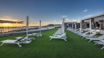Zona de lounge en la azotea con filas de tumbonas blancas y sombrillas, con vistas a un puerto deportivo con barcos y el atardecer.