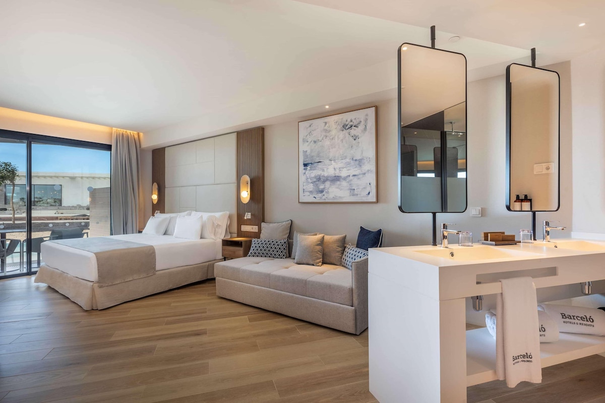 A modern hotel room with a large bed, a sofa, a desk with a mirror, and a large window offering a view.