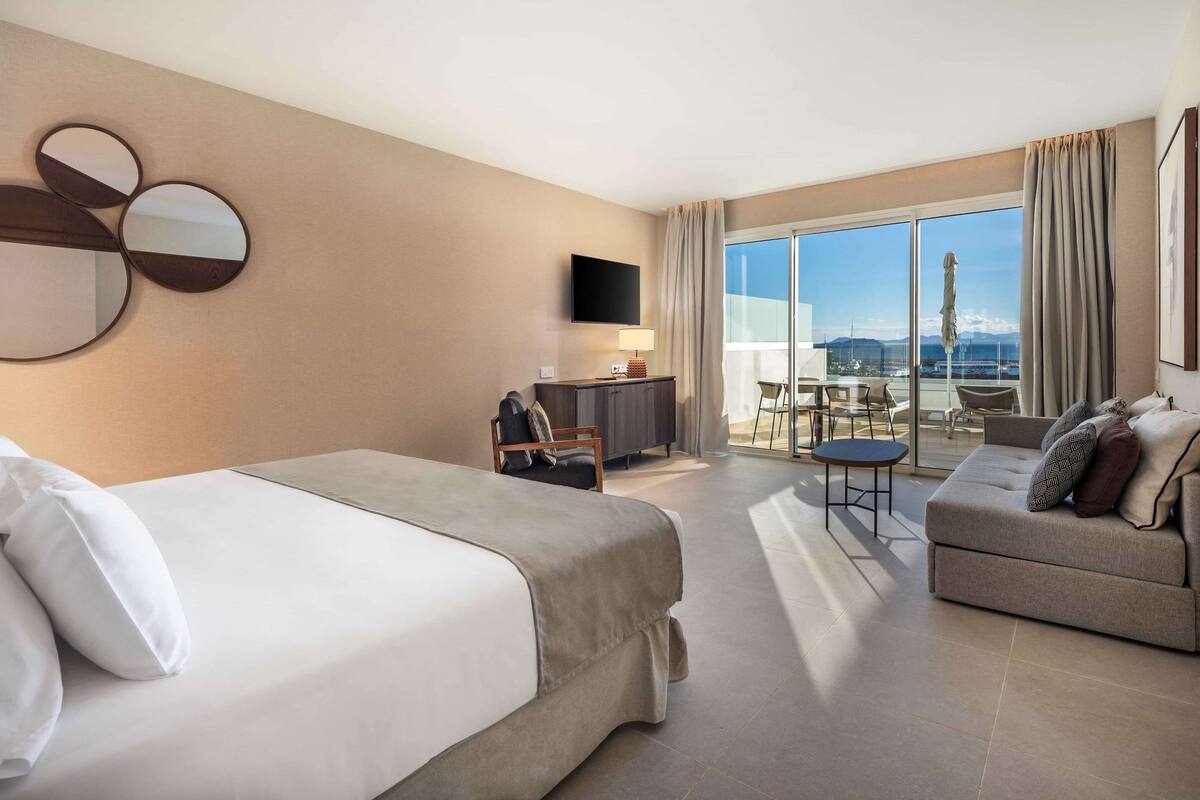 A hotel room with a bed, a sofa, a desk with a chair, a TV, and a balcony with a view.