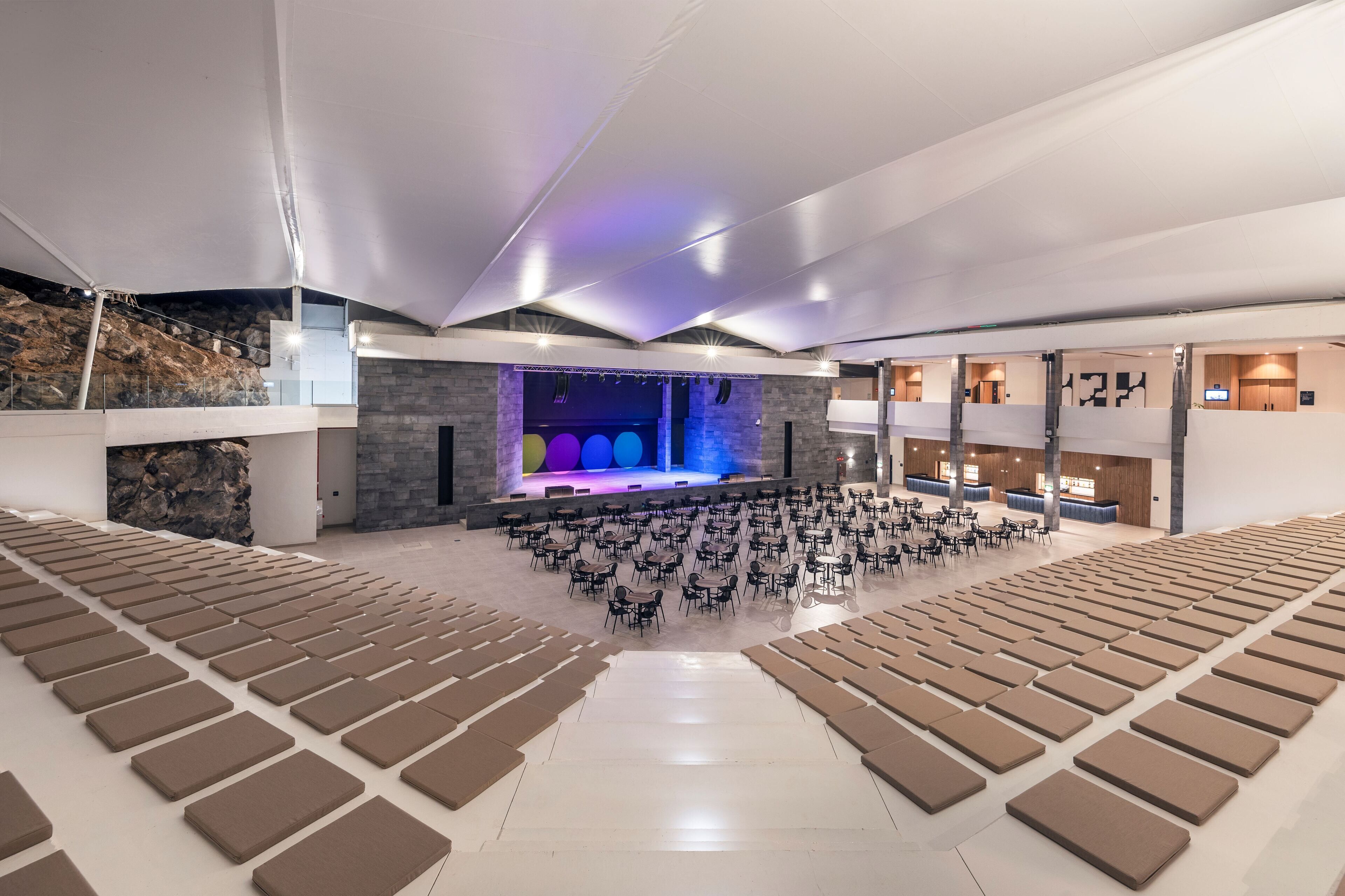 Un amplio salón con asientos escalonados, un escenario con iluminación de colores y un diseño moderno con detalles de piedra.