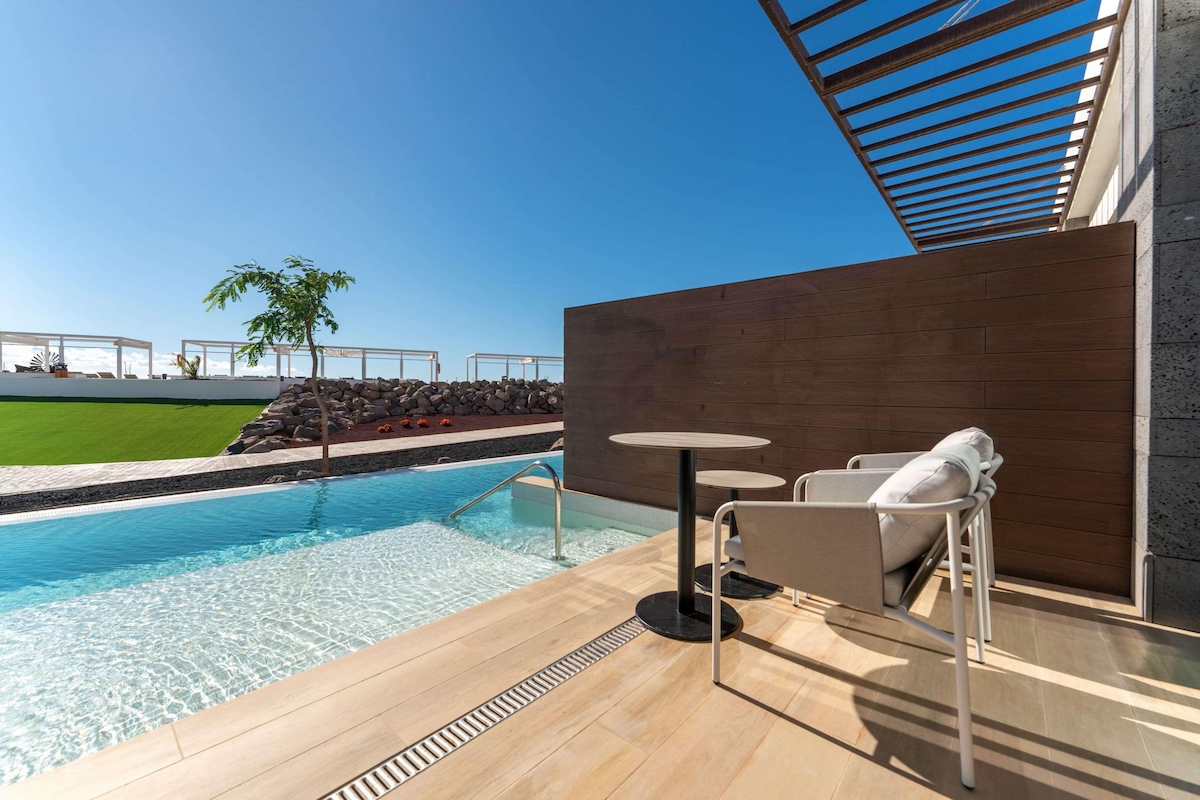 A modern outdoor pool area with a clear view of the sky, a wooden deck, and a seating area with chairs and a table.
