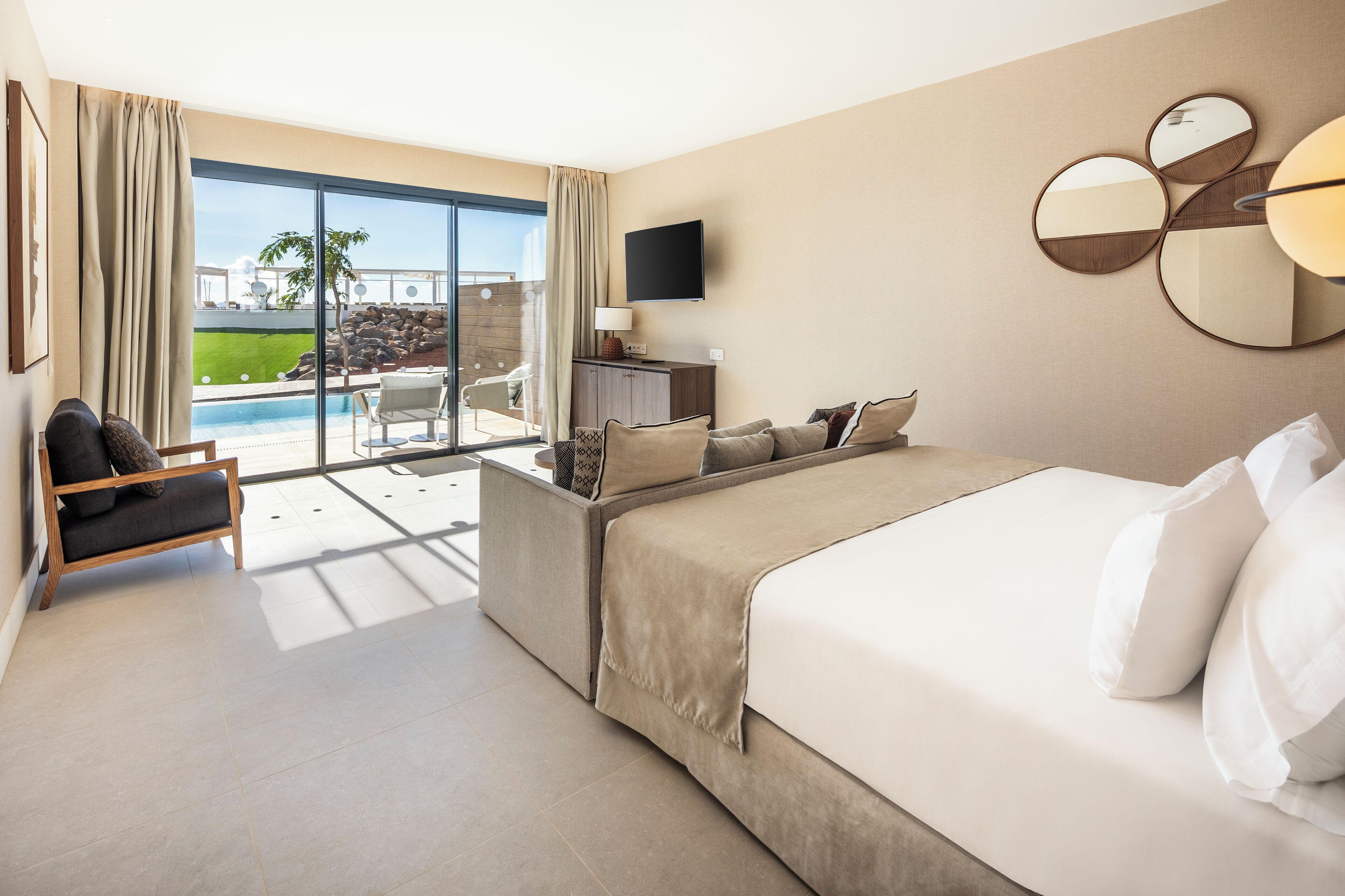 A modern hotel room with a large bed, a sofa, a TV, and a view of a swimming pool and outdoor area.