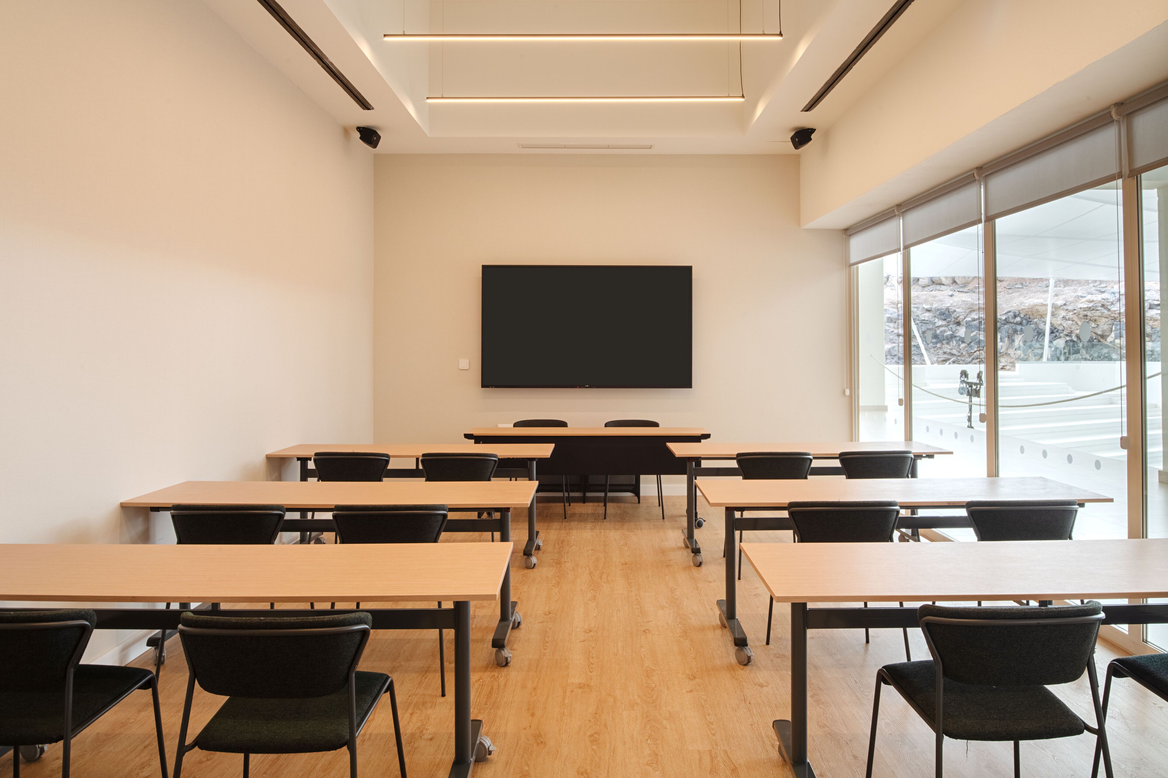 A classroom with wooden desks and black chairs, a large black screen on the wall, and large windows with a view of a rocky landscape.