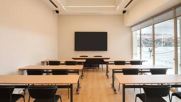 A classroom with wooden desks and black chairs, a large black screen on the wall, and large windows with a view of a rocky landscape.