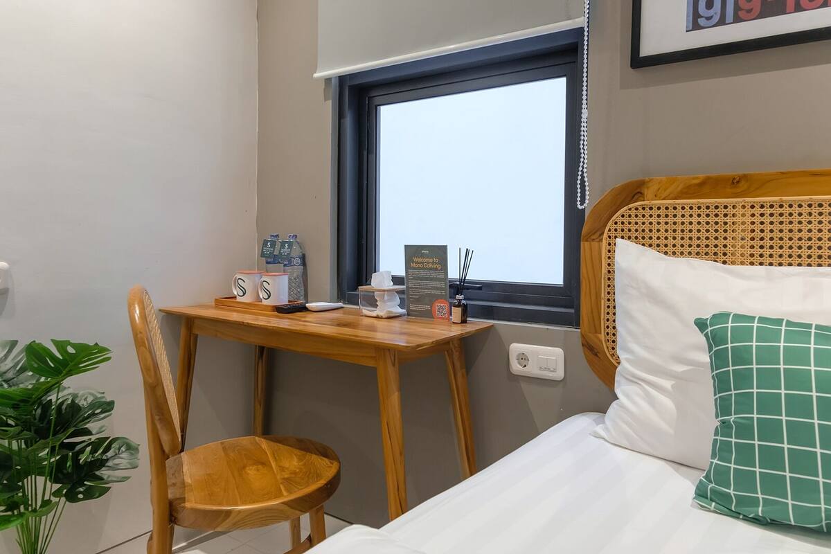 A small room with a bed, a wooden table, a chair, a window, and a potted plant.