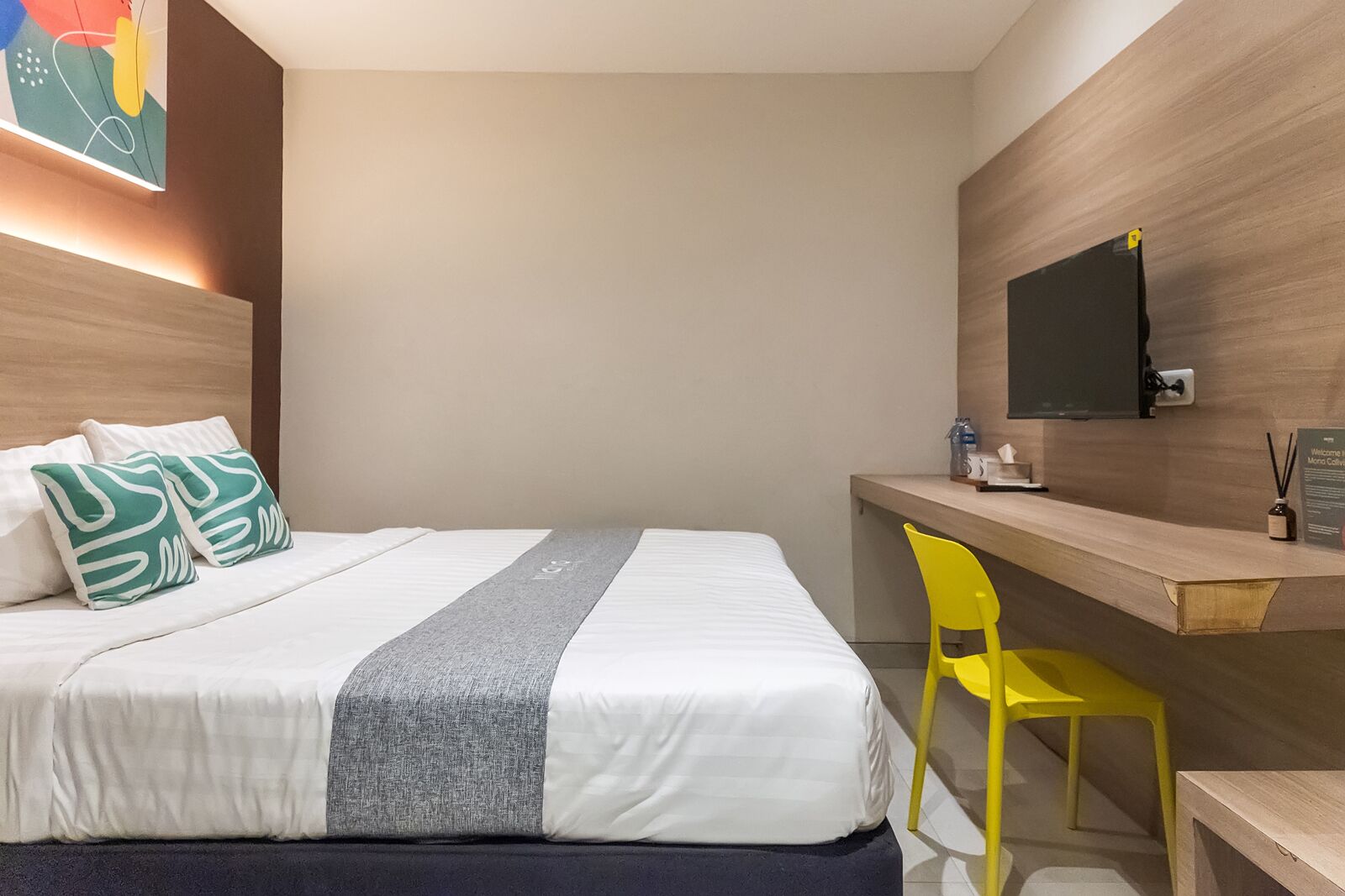 A hotel room with a bed, a desk, a chair, a TV, and a wall art piece.
