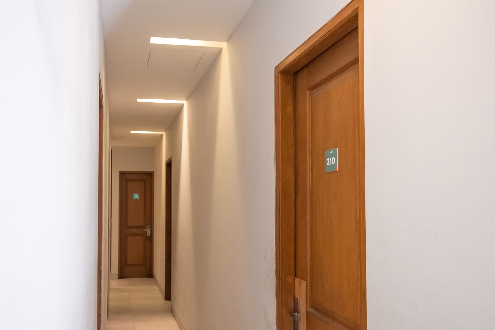 Un couloir d’hôtel avec une porte en bois portant le numéro « 210 », un plafond éclairé et un sol carrelé.