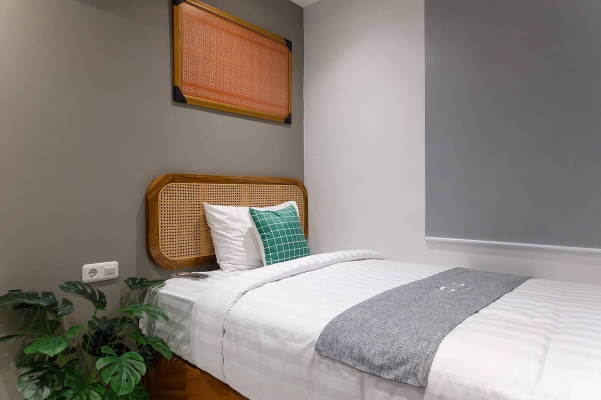 A bedroom with a rattan headboard, white bedding, a grey throw, and a green pillow.