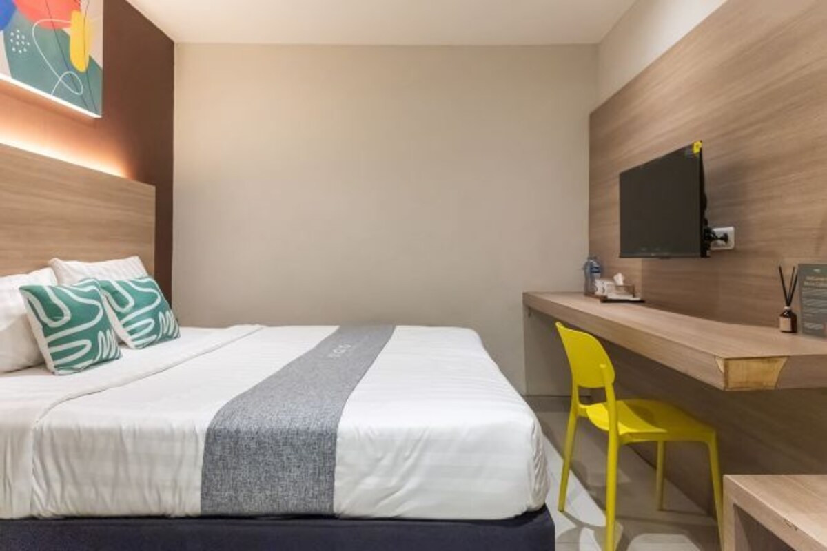 Hotelkamer met een bed, een bureau, een stoel, een aan de muur bevestigde televisie en een kleurrijk abstract schilderij boven het bed.