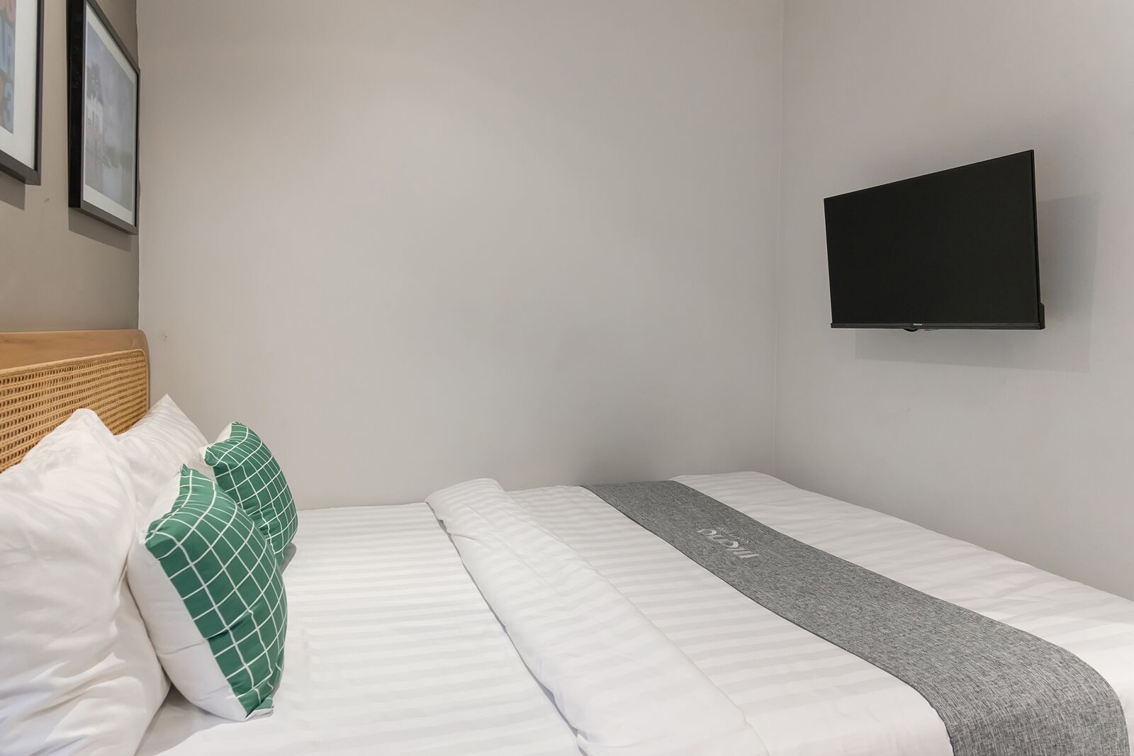 A single bed with white bedding and a gray runner, a wooden headboard, and a black TV mounted on the wall.