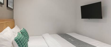 A single bed with white bedding and a gray runner, a wooden headboard, and a black TV mounted on the wall.