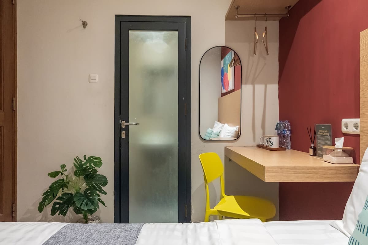 A hotel room with a bed, a desk with a chair, a mirror, a potted plant, and a door with a frosted glass panel.