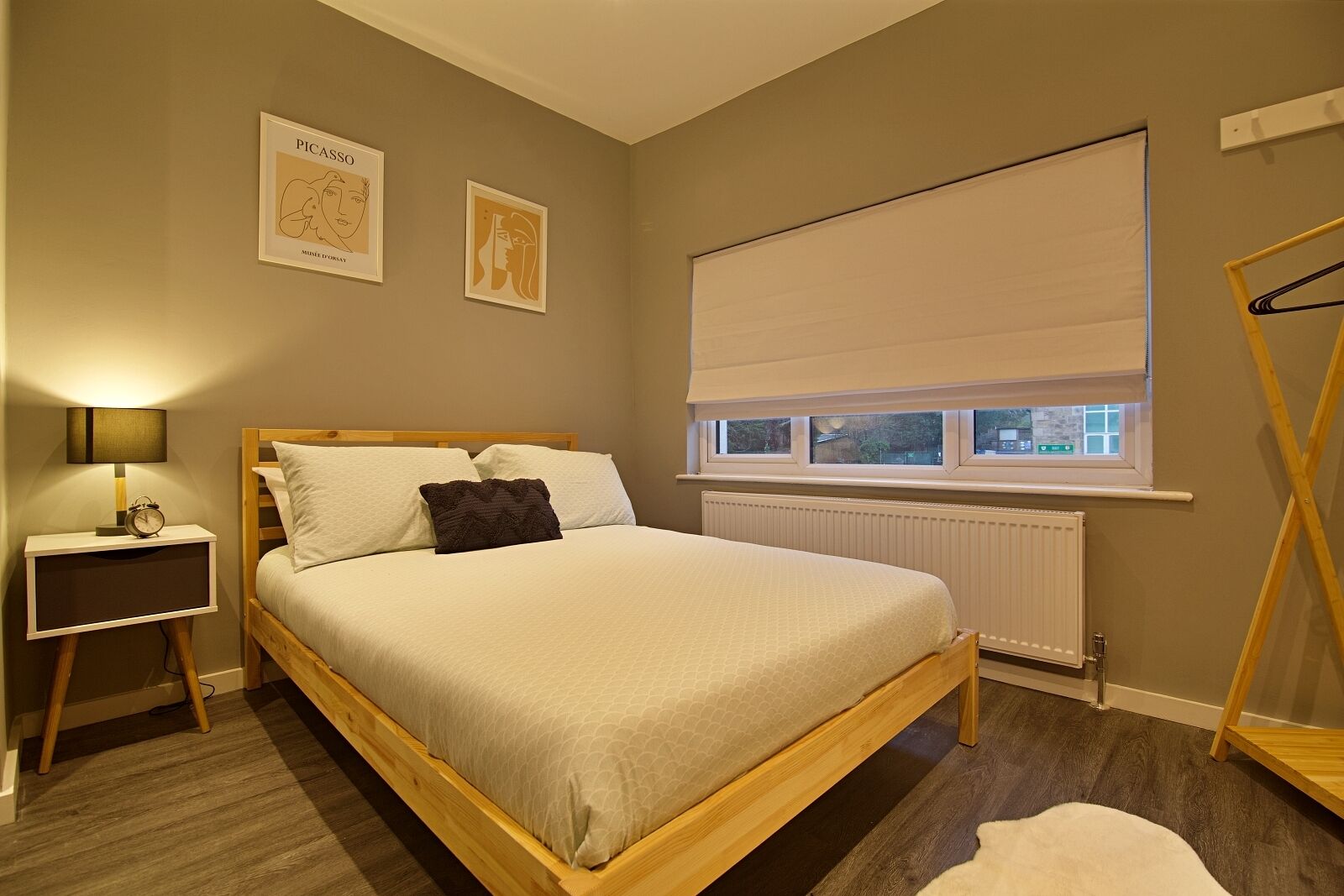 A bedroom with a wooden bed, a bedside table with a lamp, a radiator, and a window with blinds.