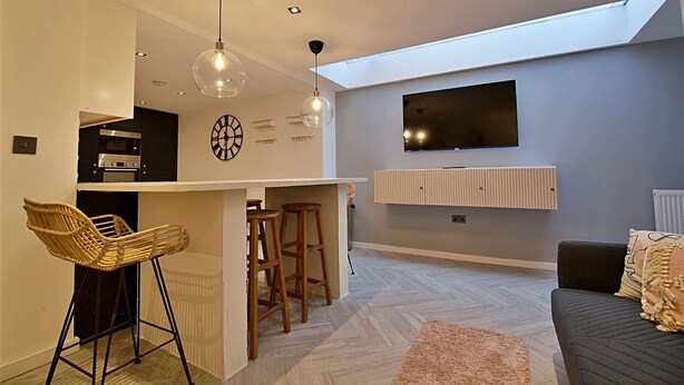 A modern kitchen with a built-in oven and microwave, a dining area with a bar stool, and a living space with a sofa and a TV mounted on the wall. - Bellerose 6 (Lancaster)