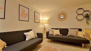 Two dark grey sofas with white and patterned cushions, a floor lamp, and wall art including framed botanical prints and geometric wall decor. - Bellerose 6 (Lancaster)