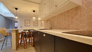 A modern kitchen with dark cabinets, a white countertop, and a hexagonal tile backsplash. There are two wooden stools and a hanging pendant light. - Bellerose 6 (Lancaster)