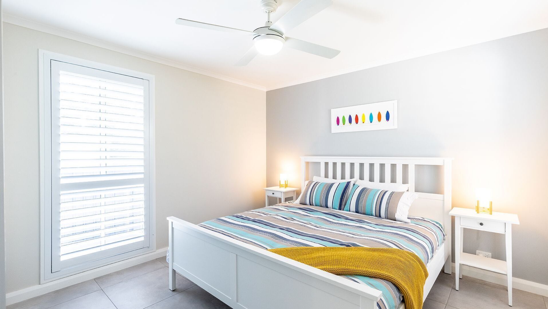 Une chambre à coucher avec un lit, des tables de chevet blanches, un ventilateur de plafond, une fenêtre avec des stores et un mur orné d’une œuvre d’art colorée.