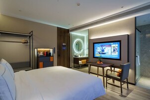 A modern hotel room with a large bed, a desk with a chair, a television, a bathroom with a mirror, and a coat rack.