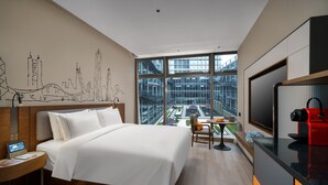 Una habitación de hotel moderna con una cama grande, un escritorio con una computadora portátil, un televisor y vistas de un paisaje urbano a través de ventanas amplias.