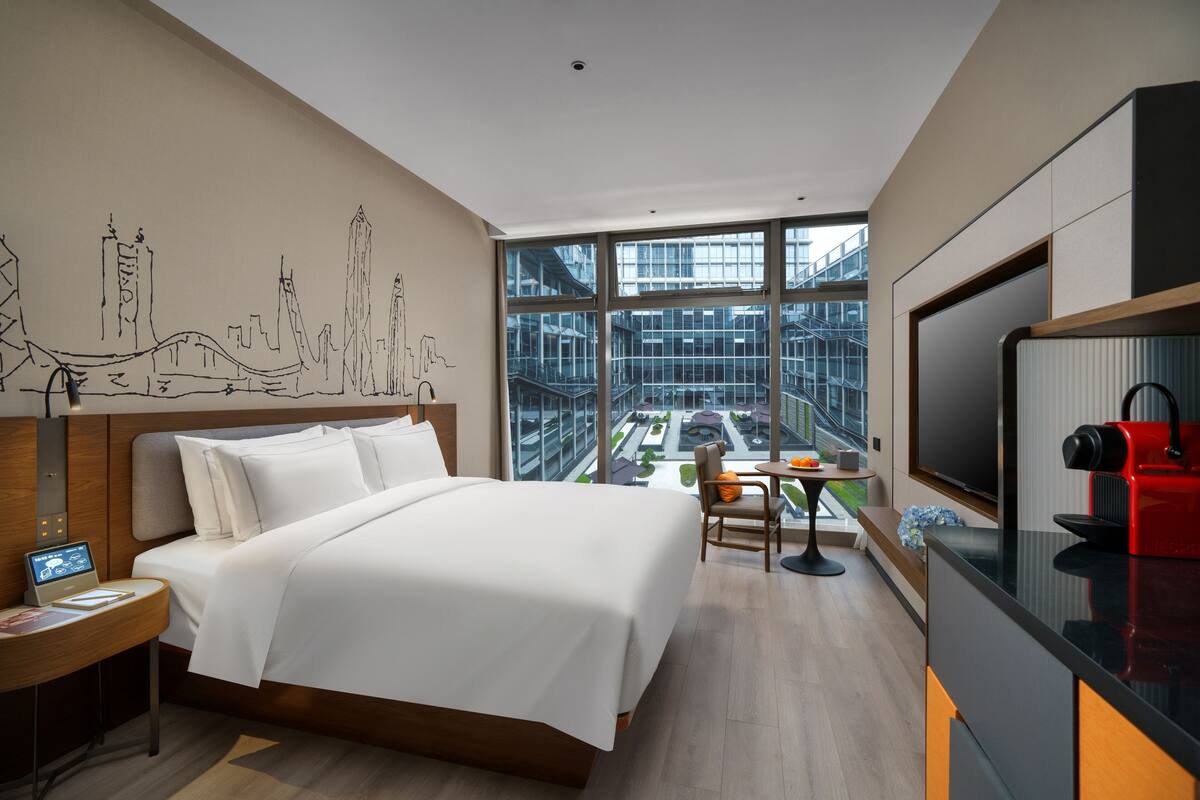 A modern hotel room with a large bed, a desk with a laptop, a TV, and a view of a cityscape through large windows.