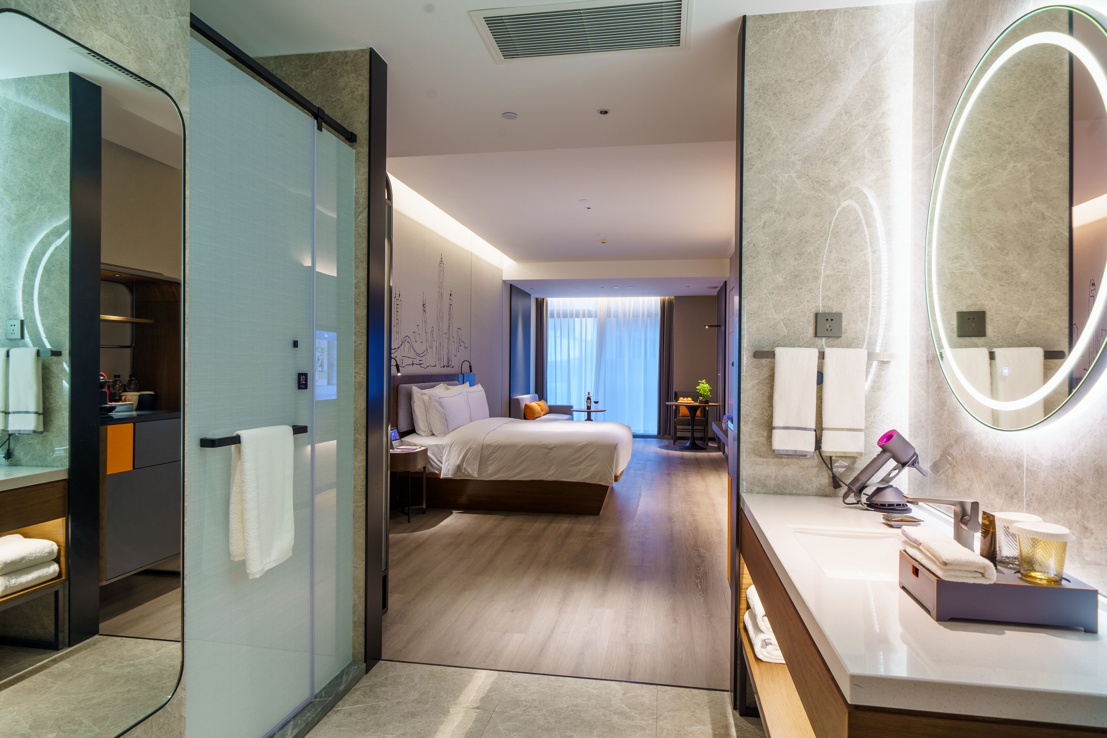 A modern hotel room with a large bed, a separate bathroom with a vanity, and a view of the cityscape through the window.
