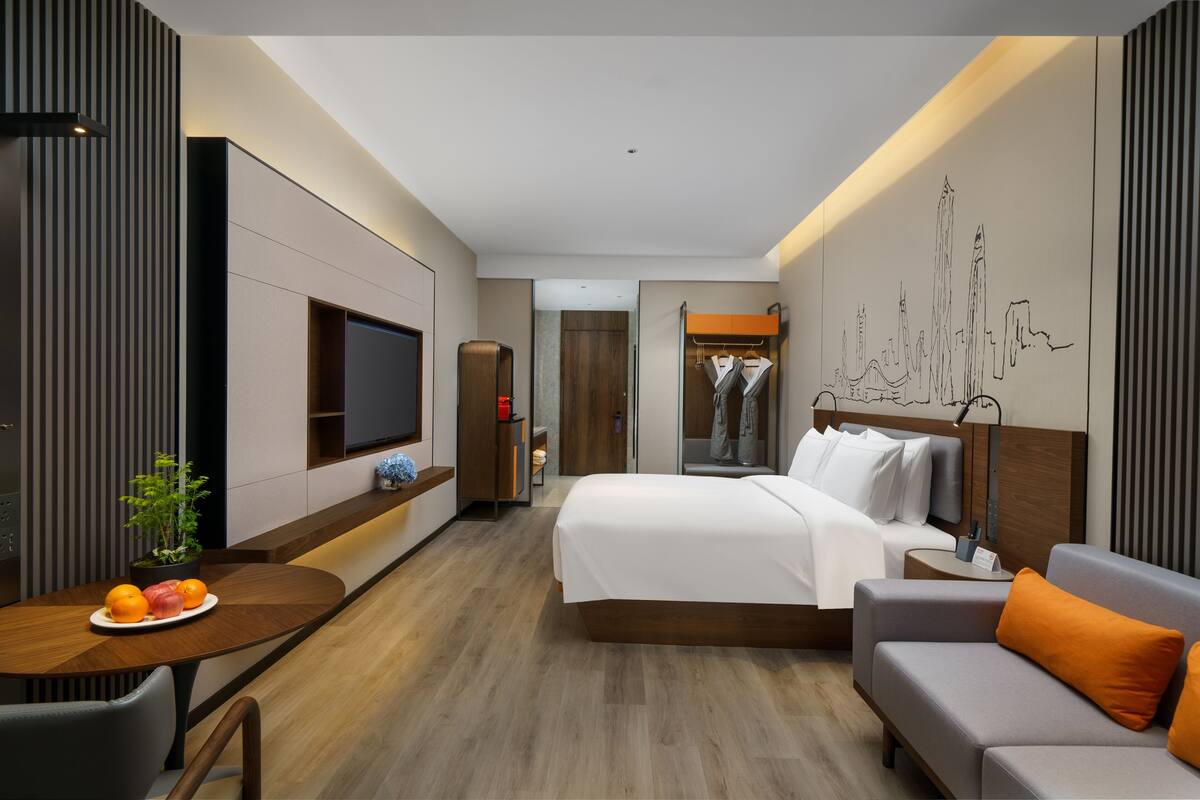 A modern hotel room with a bed, a sofa, a dining table with fruit, a TV, and a cityscape artwork on the wall.