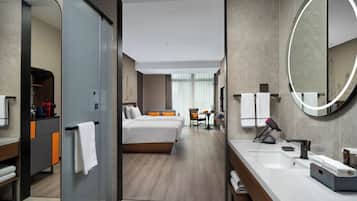 A modern hotel room with a bed, a desk, a bathroom with a mirror, and a shower area.