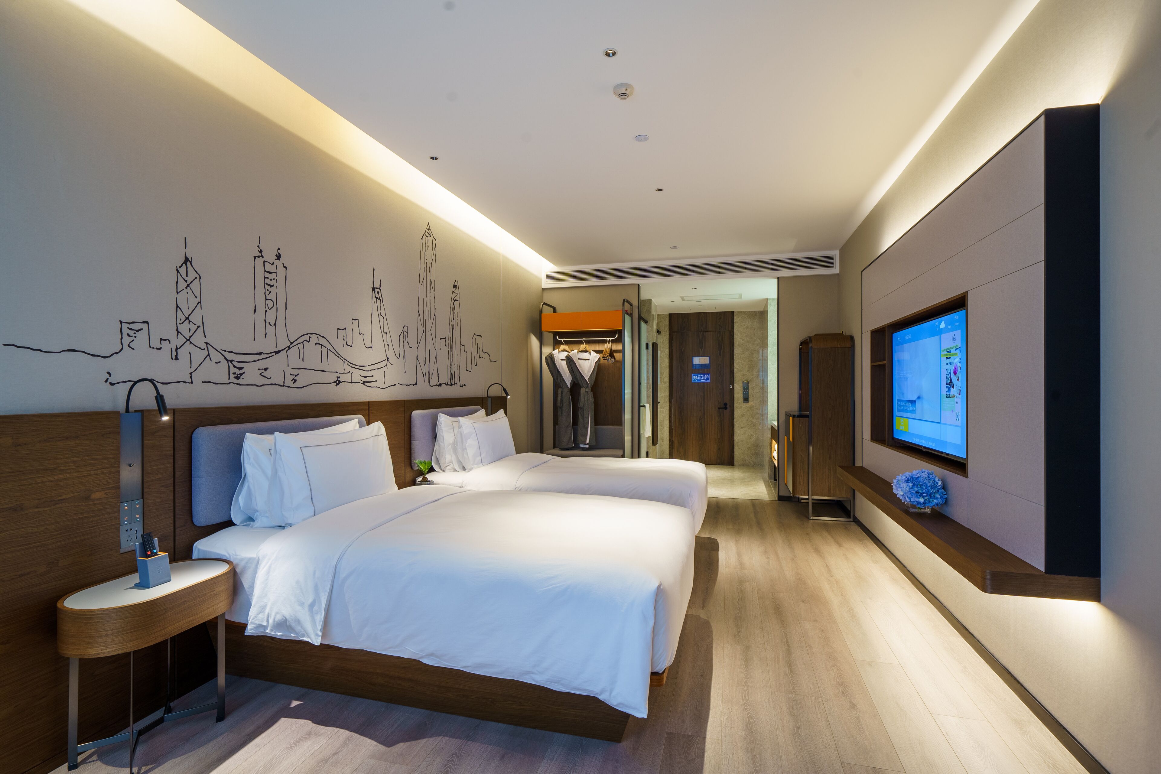 A modern hotel room with a large bed, bedside tables, a TV, and a cityscape mural on the wall.