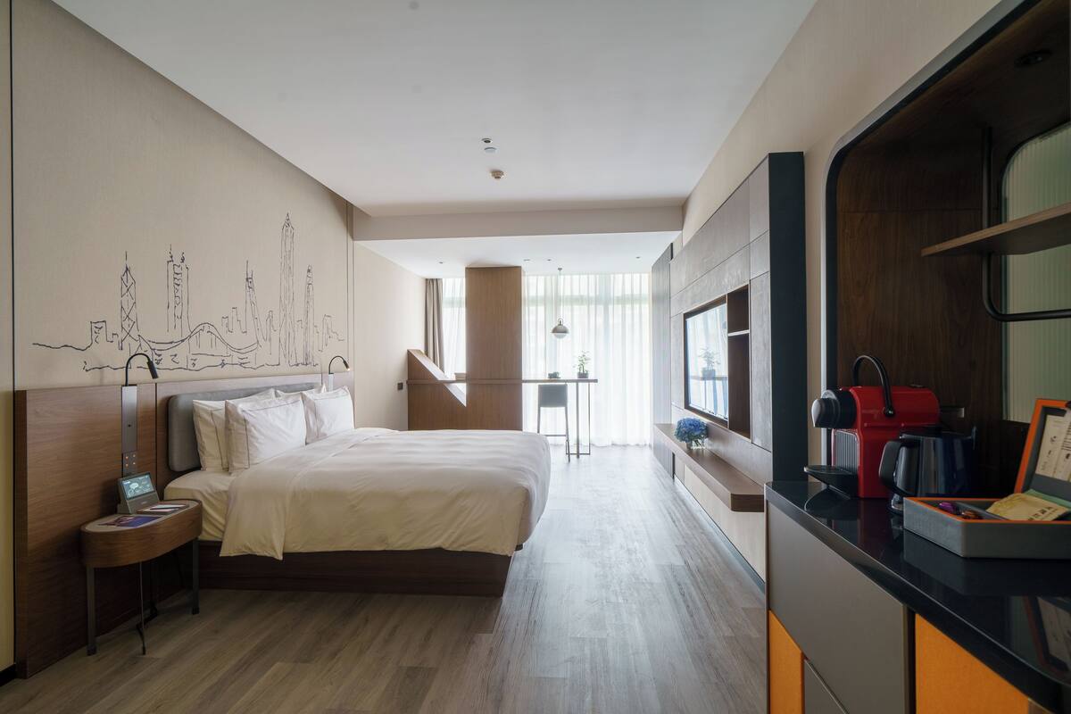 A modern hotel room with a large bed, a desk with a coffee machine, and a cityscape mural on the wall.