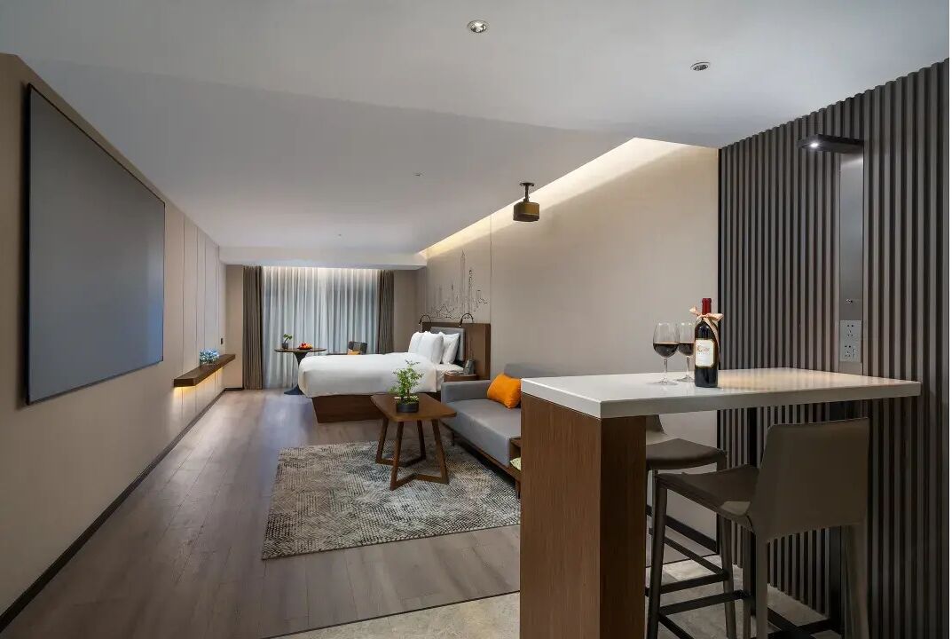 Une chambre d’hôtel moderne avec un lit, un petit coin salon, une table avec deux verres de vin et une grande fenêtre.
