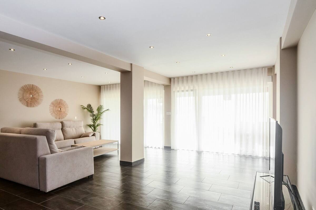 Ein modernes Wohnzimmer mit einer Couch, einem Couchtisch und einem Fernseher. Das Zimmer hat ein großes Fenster mit durchsichtigen Vorhängen und einen dunklen Holzboden.