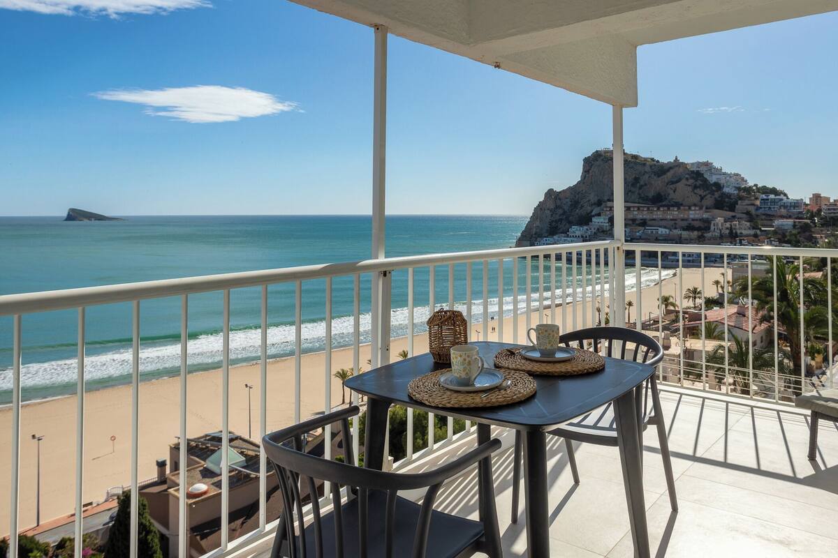 Ein Balkon mit Tisch und Stühlen mit Blick auf einen Strand und einen felsigen Hügel.