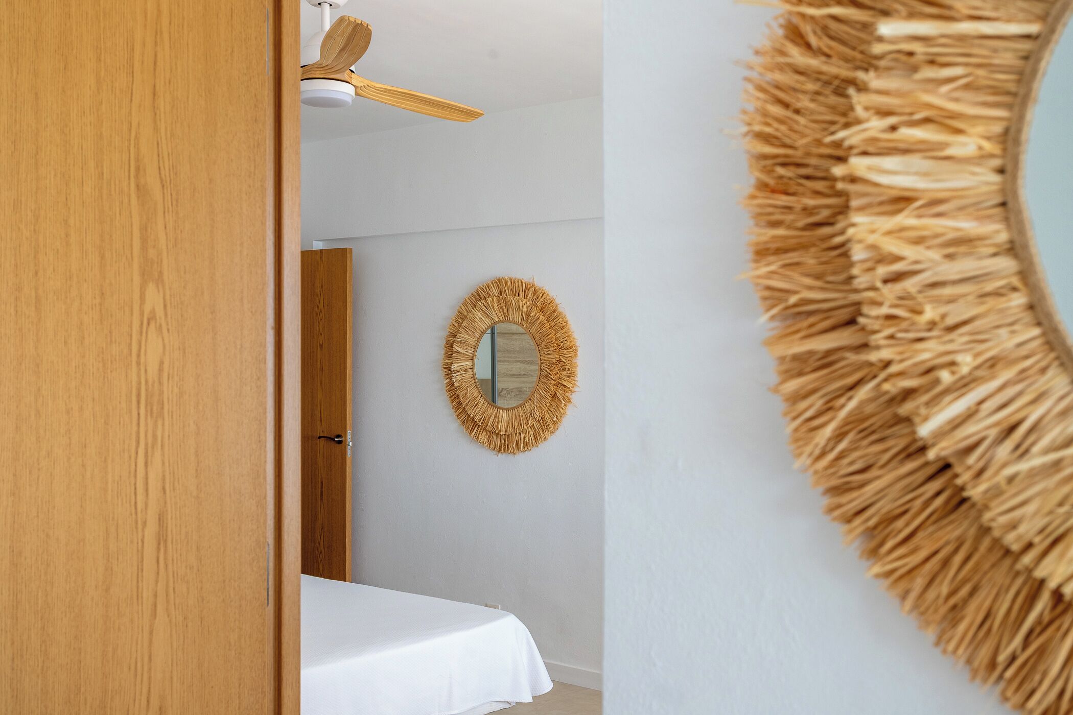 Ein Zimmer mit einem Bett, einer Holztür, einem Deckenventilator und einem wandmontierten Spiegel mit einer strohähnlichen Umrandung.