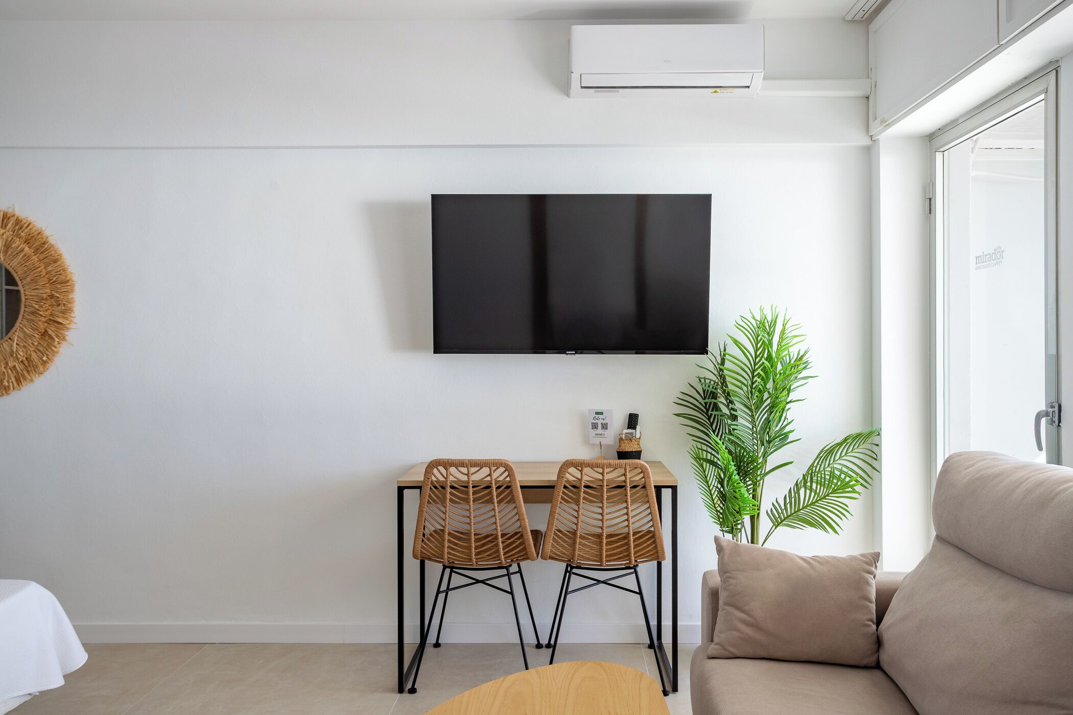 A modern living room with a flat-screen TV, two wicker chairs, a sofa, a potted plant, and a dining table.