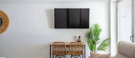 A modern living room with a flat-screen TV, two wicker chairs, a sofa, a potted plant, and a dining table.