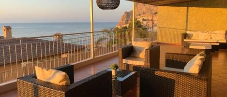 Ein Balkon mit Korbmöbeln, einer Hängelampe und Blick auf das Meer.