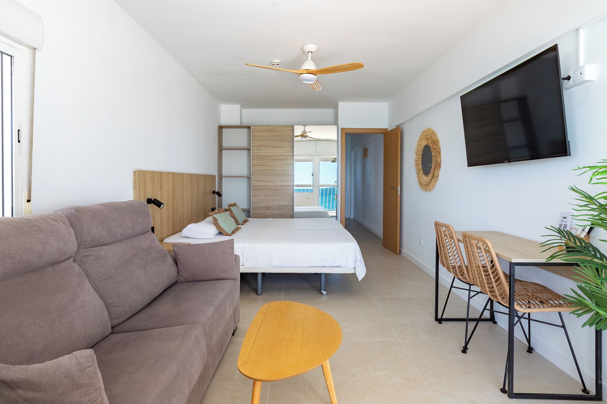A modern hotel room with a sofa, bed, dining table, chairs, TV, ceiling fan, and a view of the balcony.