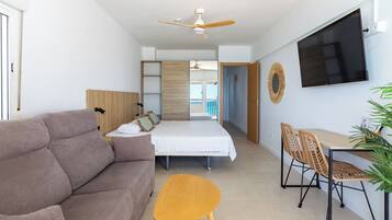 A modern hotel room with a sofa, bed, dining table, chairs, TV, ceiling fan, and a view of the balcony.