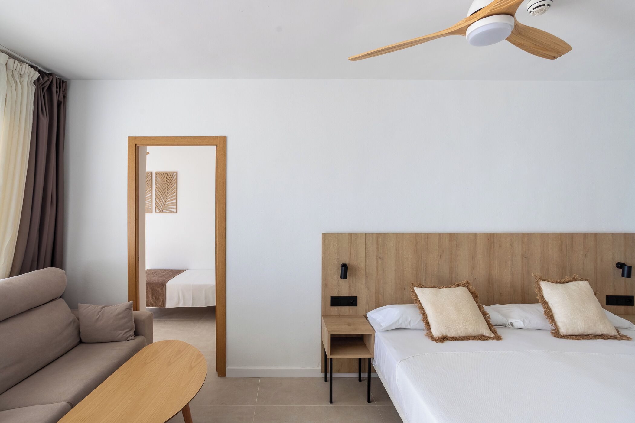 A hotel room with a bed, a wooden nightstand, a wooden table, a ceiling fan, and a view of another room through an open door.
