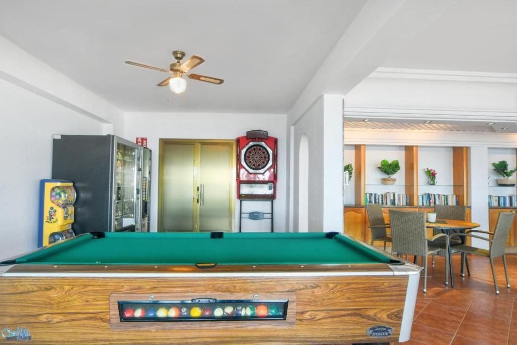 A pool table with a ceiling fan, a dartboard, and a billiards machine in a room with a dining area and bookshelves.