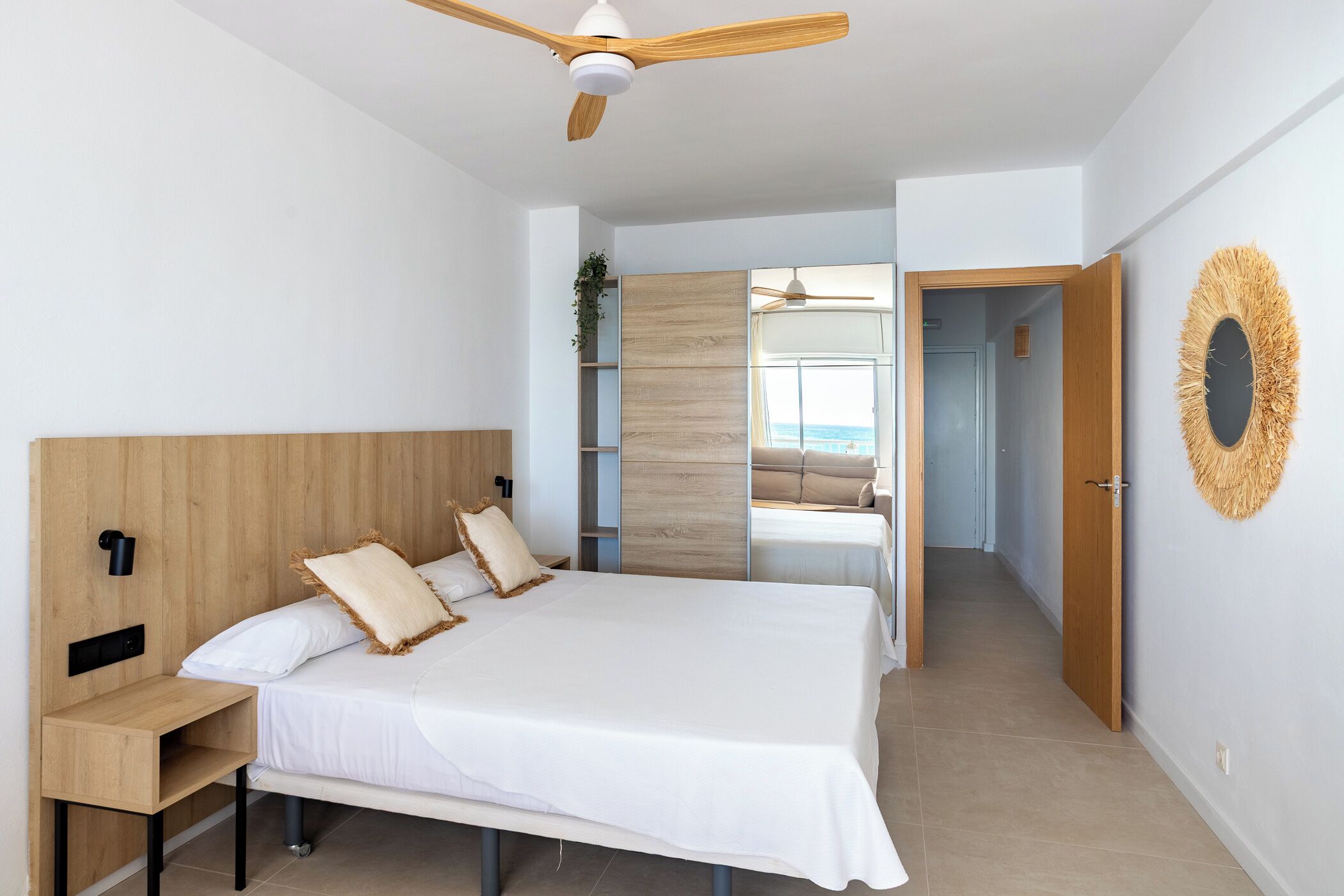 Quarto moderno com cabeceira de madeira, cama com roupa de cama branca, mesa de cabeceira, ventilador de teto e vista através de uma porta aberta.